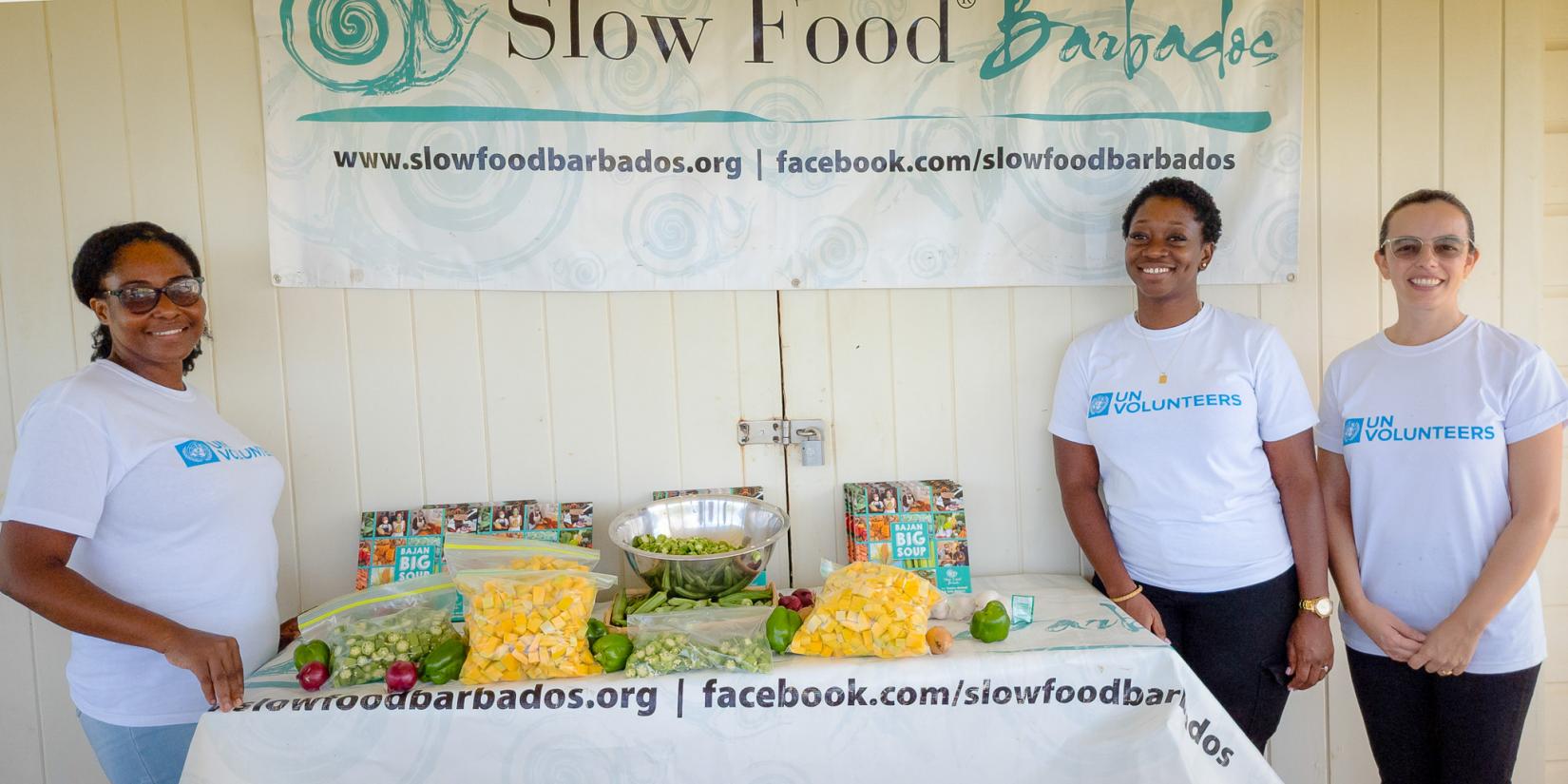 UNVs standing next to a table of freshly prepared vegetables under a Slow Food Barbados banner. 