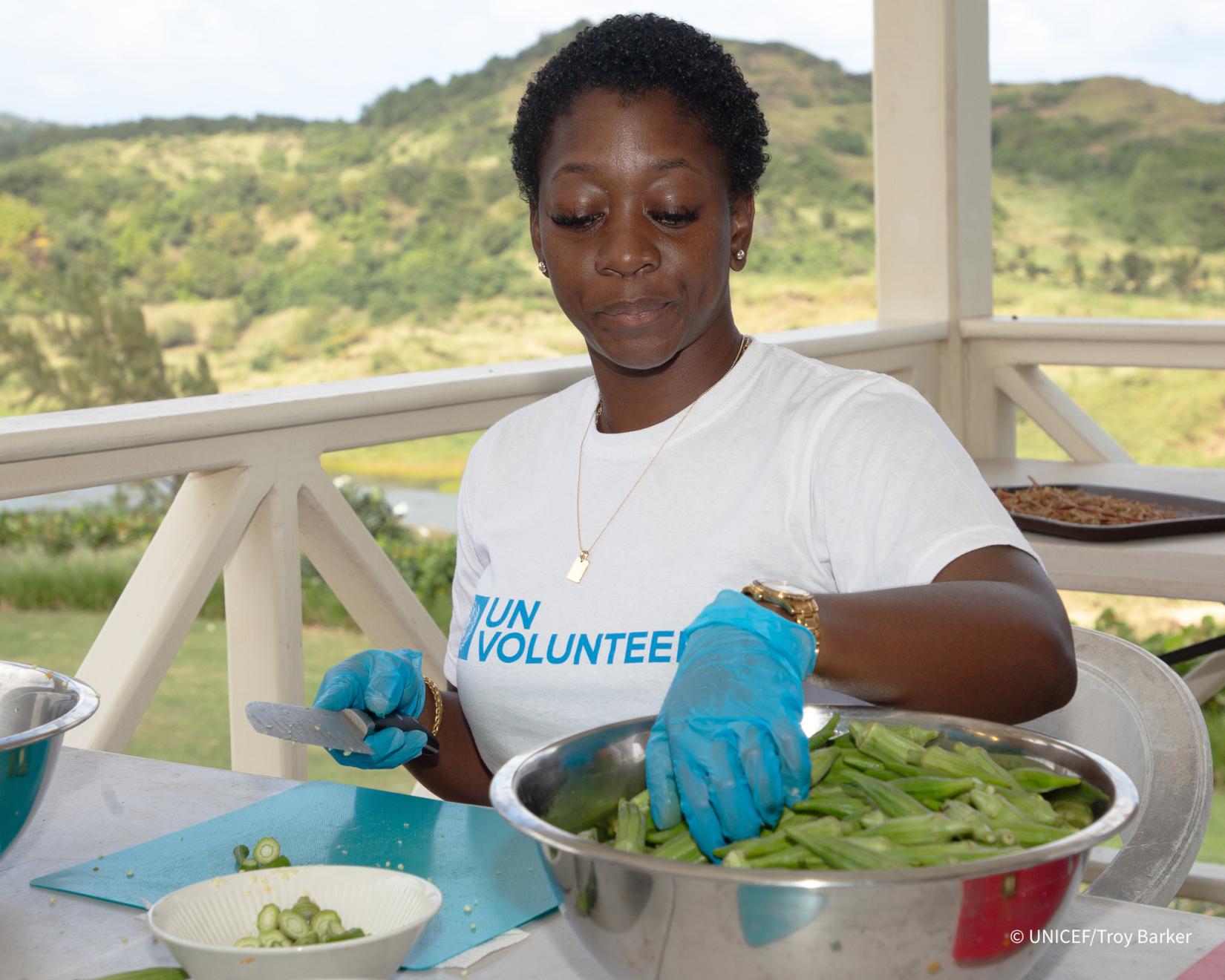 A woman picks up an okra from a bowl to cut with a knife which she is holding in the other hand while seted at a table on a balcony