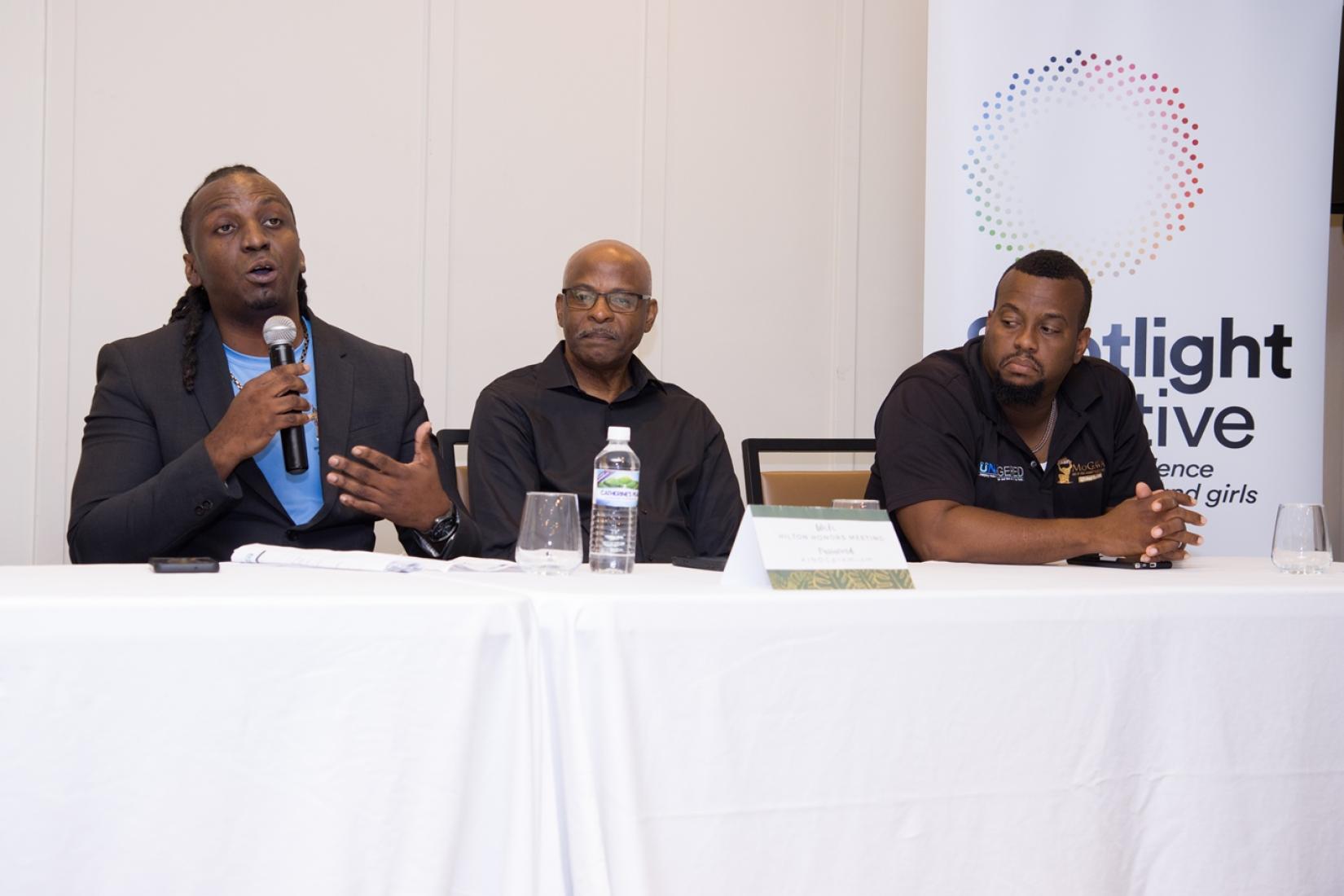 Three men sit at a table with the one on the left holding a microphone and gesturing while speaking.