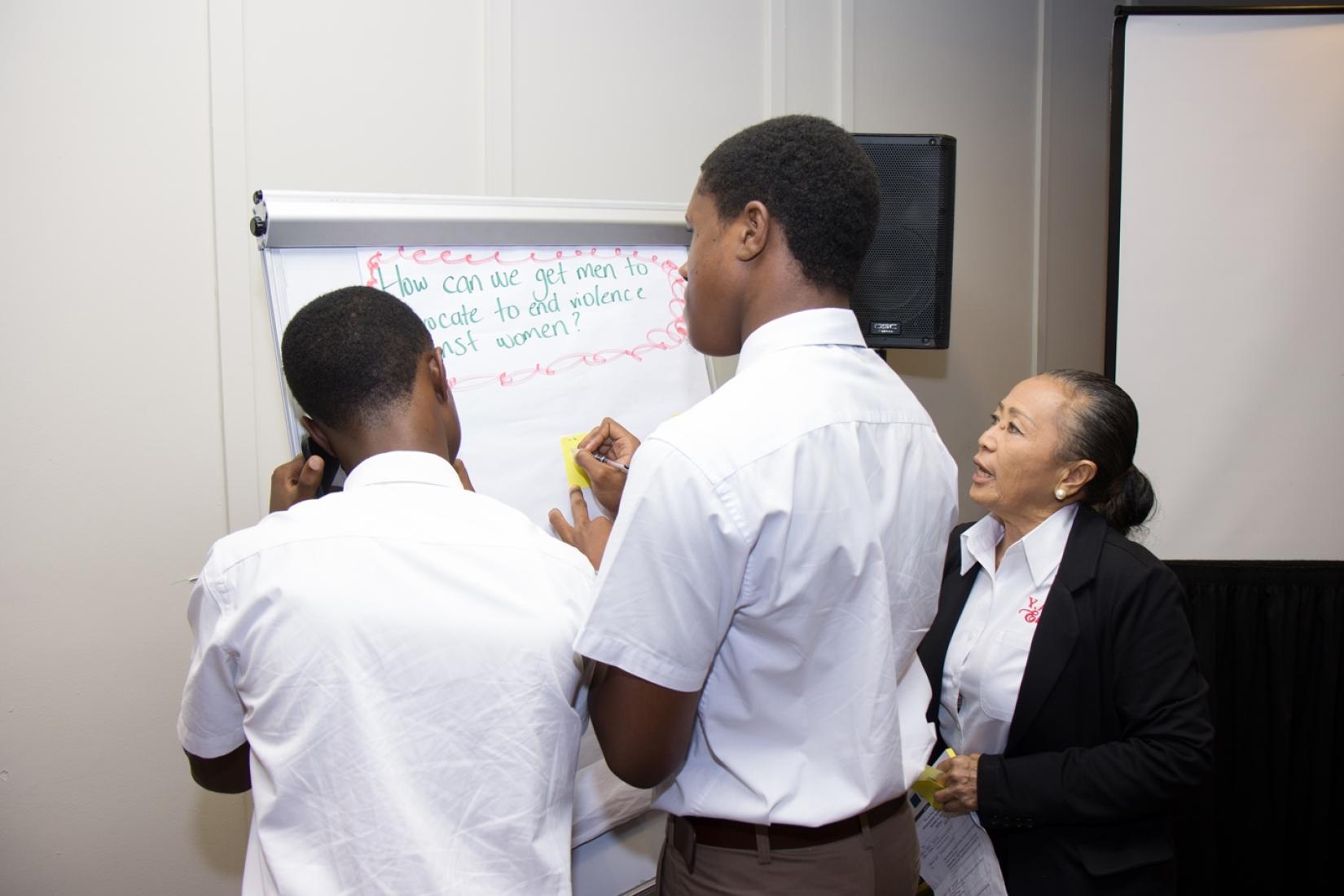 Two boys in school uniforms write on a white flip chart while a woman looks on.