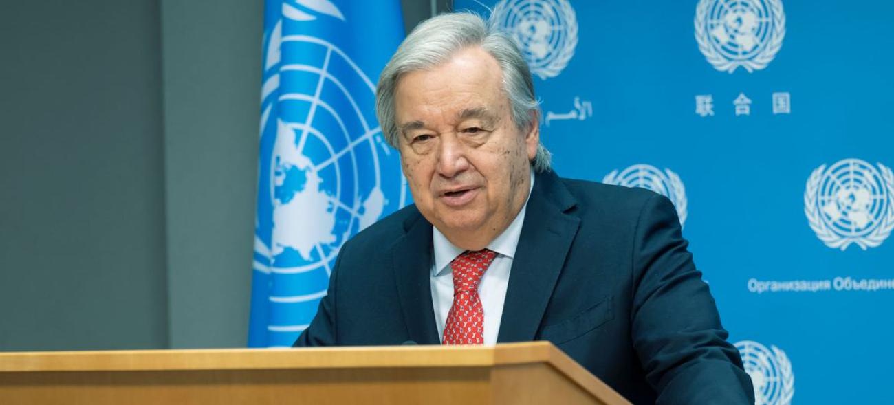 man stands at podium to speak with united Nations flag and branded backdrop behind him.