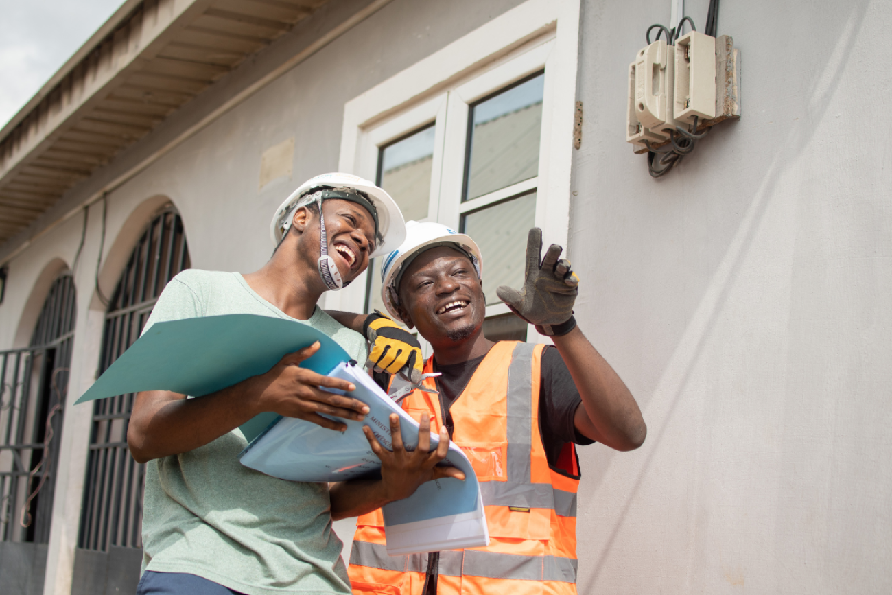 Two electricians stand together in front of a house and point at something not visible in the image