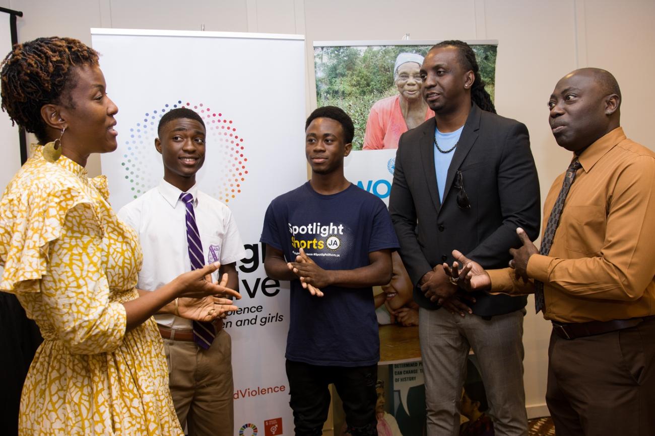 A woman and five men stand in a semi-circle having a discussion in front of promotional standing banners