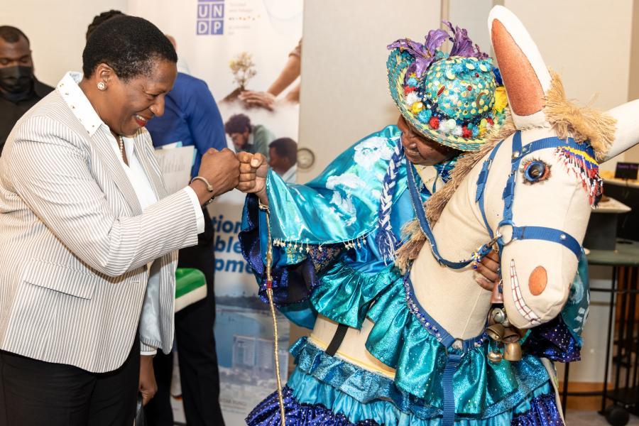 Minister of Planning and Development, Pennelope Beckles, greets a Burrokeet, a traditional mas character. The character portrayal was part of the UNDP's exhibition booth at the launch of the 2022 Annual Report Launch.