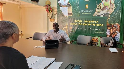 Woman interviews a man in an office setting with a backdrop highlighting Saint Lucia's cultural heritage.